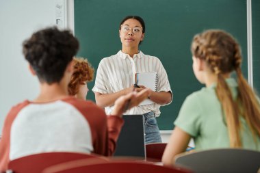 Sınıfta öğrencilerin ve dizüstü bilgisayarların yanında not defteri tutan dalgın bir Afro-Amerikan öğretmeni.