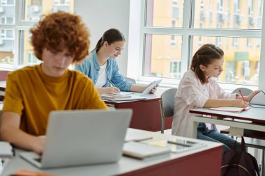 Smiling teen schoolgirl using digital tablet and writing on notebook near classmates in school clipart