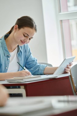 Sınıfta ders sırasında dijital tablet kullanan ve deftere yazı yazan gülümseyen genç öğrenci.