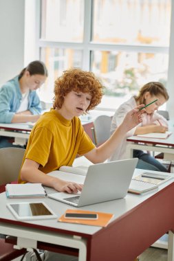 Redhead teen schoolboy holding pencil and using laptop while talking during lesson in classroom clipart