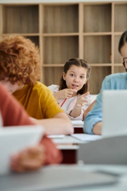 Öğrenci kız ders sırasında parmak ucunda sınıf arkadaşlarının yanında konuşuyor ve işaret ediyor.