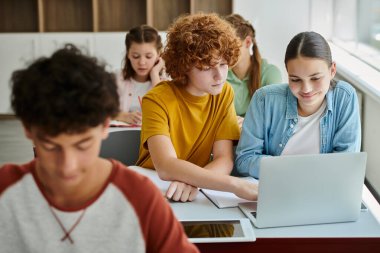 Redhead schoolboy using laptop with smiling classmate together during lesson in blurred school clipart