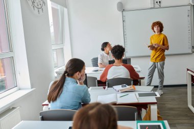 Elinde kitap tutan ve ders sırasında Afrika kökenli Amerikalı öğretmen ve arkadaşlarının yanında duran genç bir öğrenci.