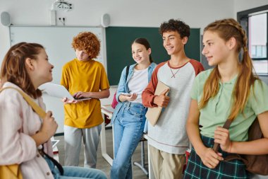Positive teenage pupils with devices and backpacks talking while standing in classroom in school clipart