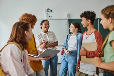 happy african american teacher talking to teenage students in classroom, back to school concept clipart