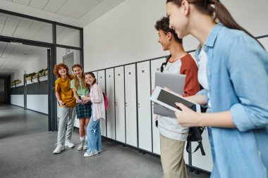 happy students looking at teen classmates with devices in school hallway, back to school concept clipart