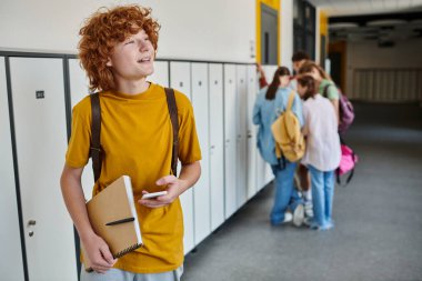 Akıllı telefon kullanan, elinde not defteri, bulanık, okul koridorunda öğrencileri tutan neşeli kızıl okul çocuğu.