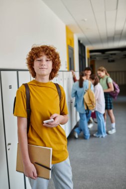 redhead schoolboy holding smartphone, happy student in school hallway looking at camera during break clipart