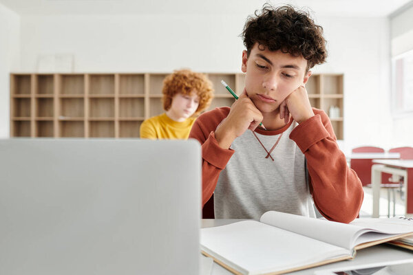 Pensive teen schoolboy looking at notebook near laptop and digital tablet in school classroom