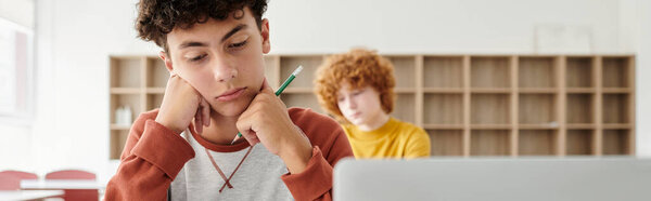 Pensive teen schoolboy holding pencil and looking at laptop near blurred friend in classroom, banner
