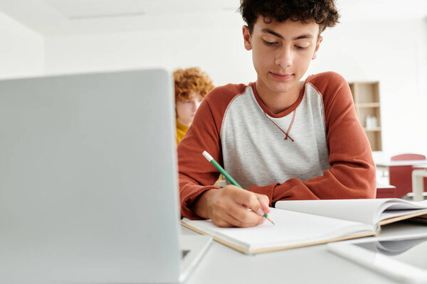Teenage schoolboy writing on notebook near laptop and blurred classmate in classroom in school