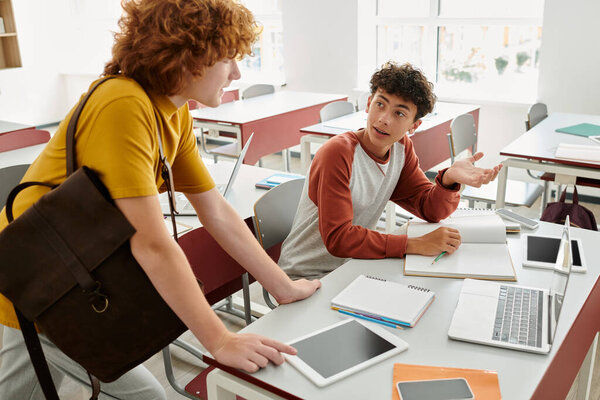 Teenage schoolboy talking to friend with backpack near devices and notebooks on desk in classroom
