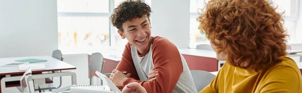 Stock image Smiling teen schoolboy talking to classmate near devices during lesson in classroom, banner