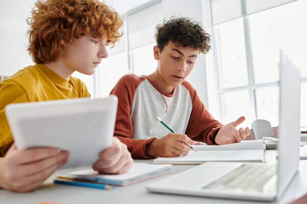Teenage schoolboy writing on notebook near redhead friend with digital tablet in classroom
