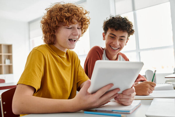 Positive teenage schoolboy using digital tablet with friend during lesson in classroom at background