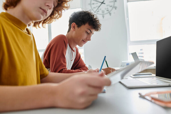 Teenage schoolboy writing while sitting near classmate and devices in classroom at background