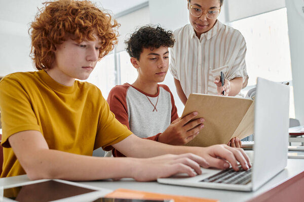 African american teacher standing near teen schoolboys and devices during lesson in school