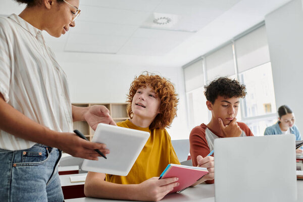 Teen schoolboy talking to african american teacher during lesson with devices in classroom