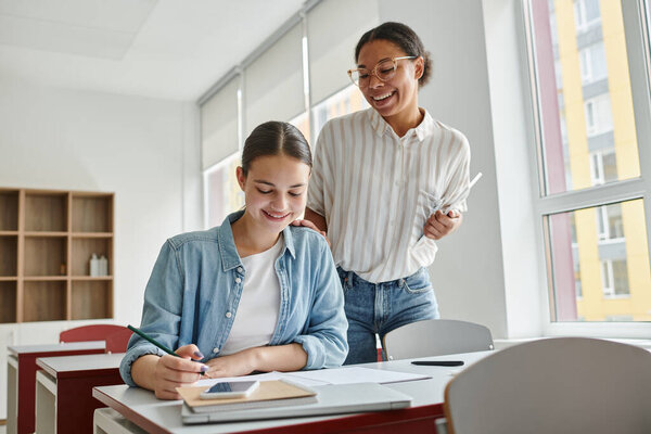 Happy african american teacher with digital tablet standing near schoolgirl during lesson in school
