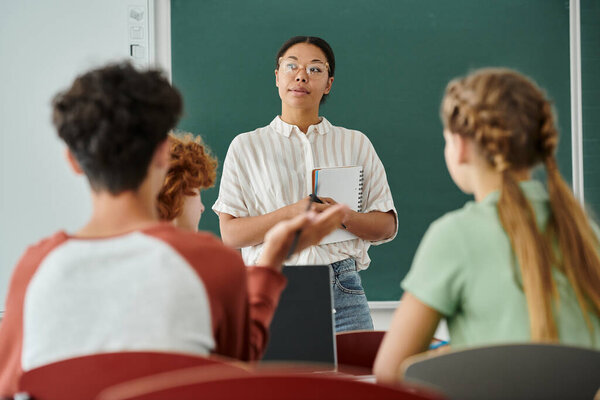 Pensive african american teacher holding notebook near pupils and laptop in classroom