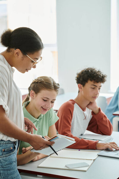 African american teacher holding notebook near positive schoolgirl during lesson in classroom