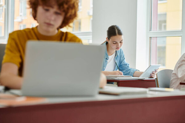 Teen schoolgirl holding digital tablet and writing on notebook near blurred classmate in classroom