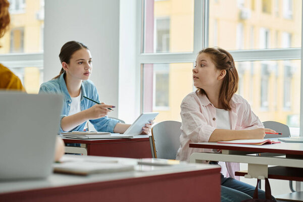 Teen schoolgirls talking while using devices and notebooks during lesson in classroom in school