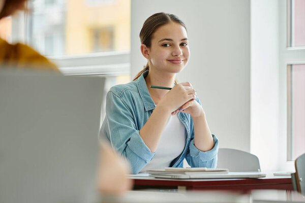 Smiling teenage schoolgirl looking at camera and holding pen near devices during lesson in classroom