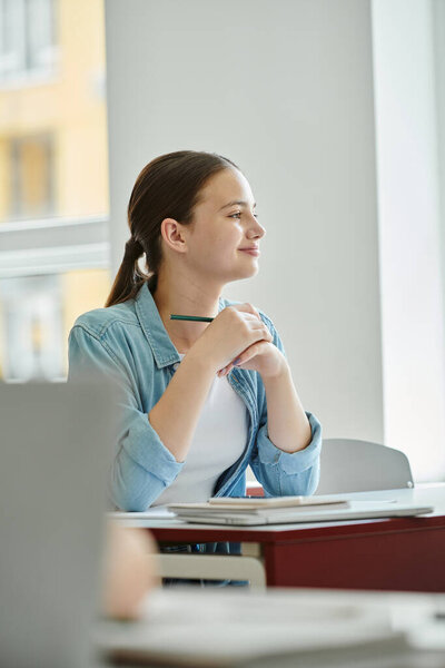 Smiling teenage schoolgirl holding pen and looking away near devices in classroom in school