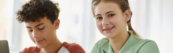 Smiling teen schoolgirl looking at camera near blurred classmate in school, banner