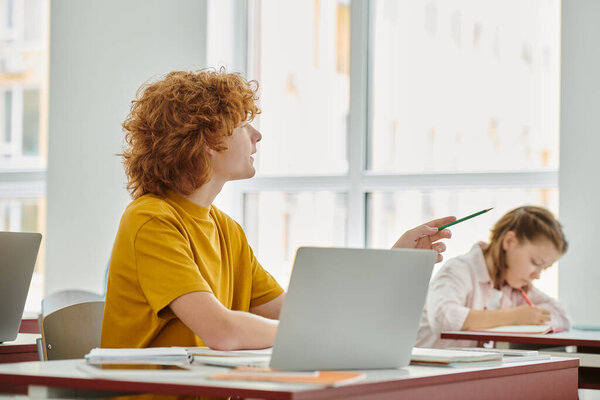 Teen redhead schoolboy talking near laptop and blurred classmate during lesson in classroom
