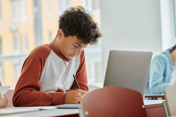 Curly teenage schoolboy writing on notebook near laptop during lesson in classroom in school