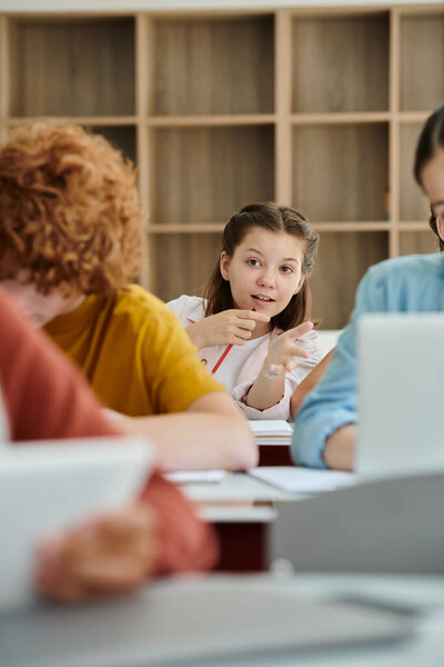 Teenage schoolgirl talking and pointing with finger near blurred classmates during lesson in class