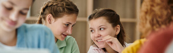 Smiling schoolgirl talking to teenage friend during lesson in blurred class in school, banner