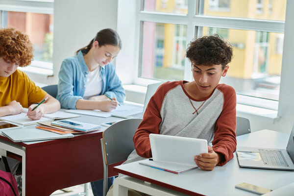 Teenage schoolboy using digital tablet near devices with blank screen and classmates in classroom