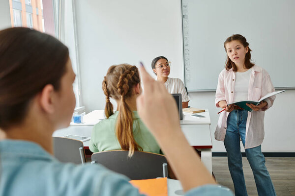 Teen schoolgirl holding notebook near classmate raising hand and african american teacher in class
