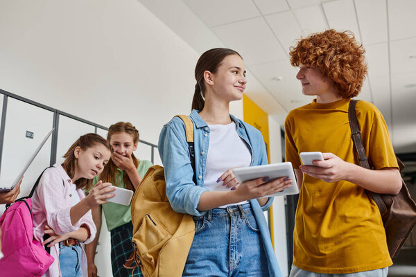 счастливый мальчик и девочка, держа гаджеты возле одноклассников в школьном коридоре, обратно к школьной концепции