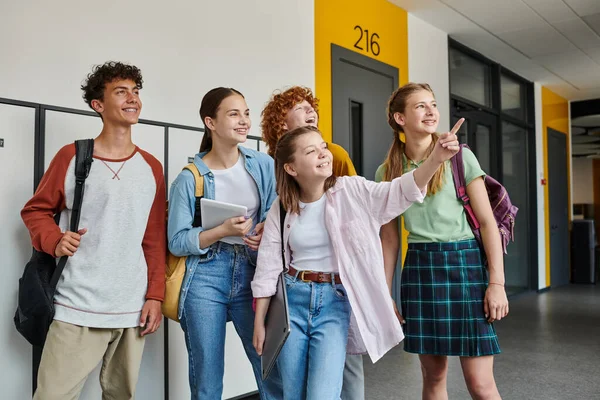 happy teen students looking away in school hallway, schoolgirl pointing with finger away, classmates