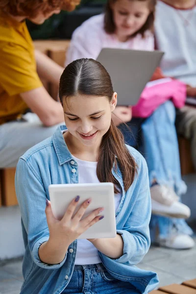 stock image back to school, teen girl using gadget near blurred classmates outdoors, break, e-study, technology