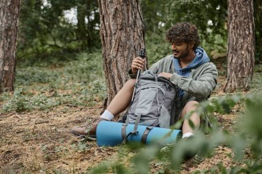 pozitif genç Hintli yürüyüşçü ormanda sırt çantasından sırık alıyor.