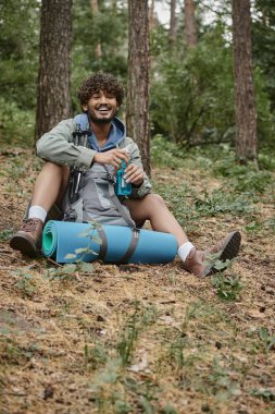 Gamsız Hintli yürüyüşçü sırt çantasının yanında spor şişesi tutuyor ve ormanda yürüyüş direkleri taşıyor.