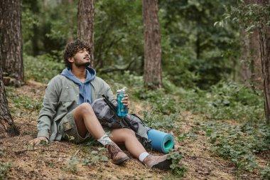 Rahat genç Hintli turist ormanda yerde otururken sırt çantasının yanında spor şişesi tutuyor.