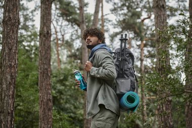 Genç kıvırcık Hintli turist ormanda sırt çantası ve spor şişesi tutuyor.