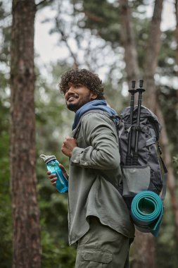 pozitif genç Hintli gezgin ormanda yürürken sırt çantası ve spor şişesi tutuyor.