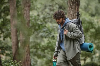 young indian tourist with backpack, trekking poles and sports bottle walking in forest clipart