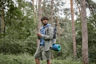 Sırt çantalı genç Hintli yürüyüşçü ormanda dururken akıllı telefon kullanıyor.