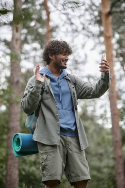 Sırt çantalı neşeli Hintli yürüyüşçü bulanık ormandaki akıllı telefondan video çağrısı alıyor.