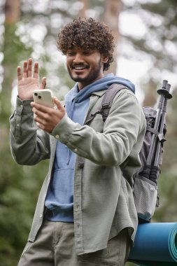 overjoyed indian hiker with backpack waving hand while having video call on smartphone in forest clipart