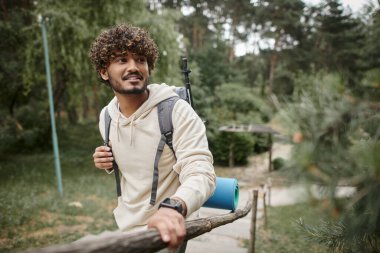 Sırt çantası ve akıllı bacaklı gülümseyen Hintli turist ormanda çitin yanında duruyor.