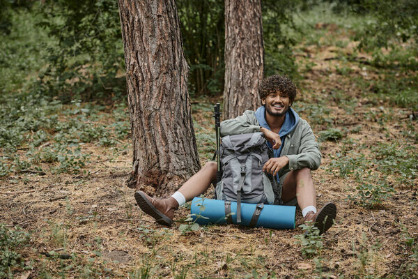 cheerful young indian man looking at camera near backpack on ground in forest